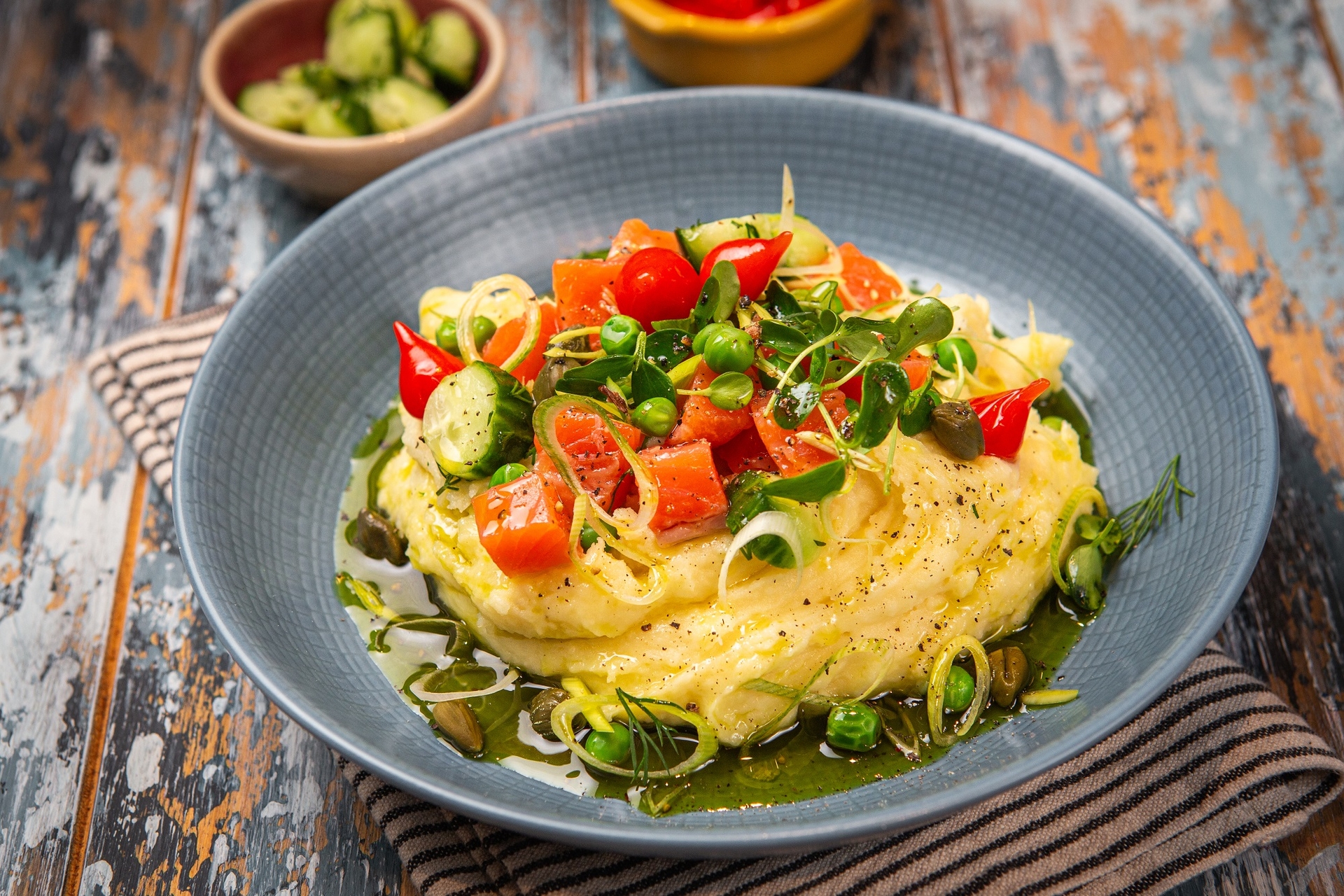 puree ziemniaczane podane w miseczce w restauracji, posypane łososiem pokrojonym w kostkę, papryczkami drop pepper i ogórkiem, polane zieloną oliwą koperkową